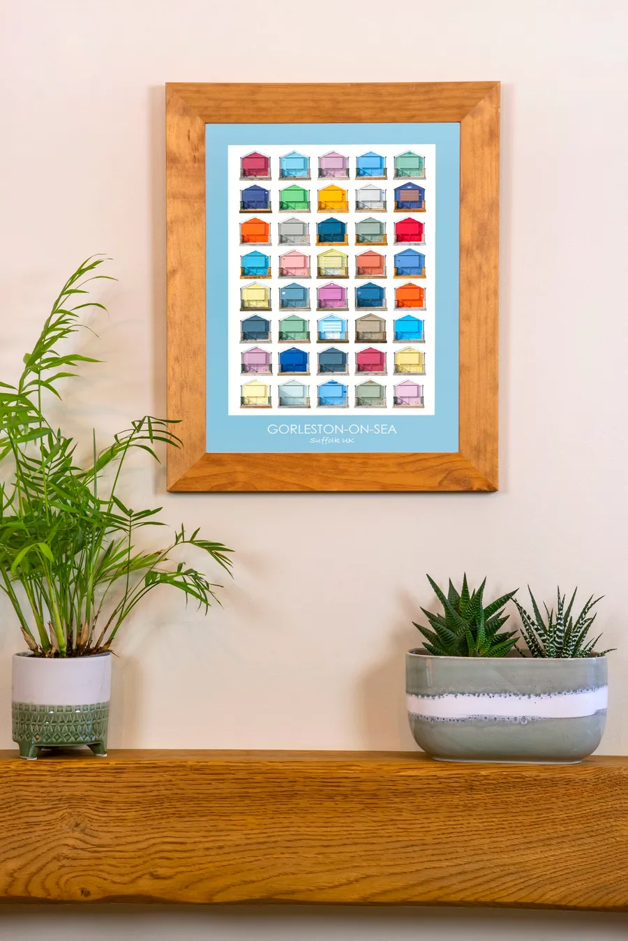The Gorleston-on-Sea print with a sky blue border in an oak frame on a cream wall above a wooden mantelpiece, between two potted plants