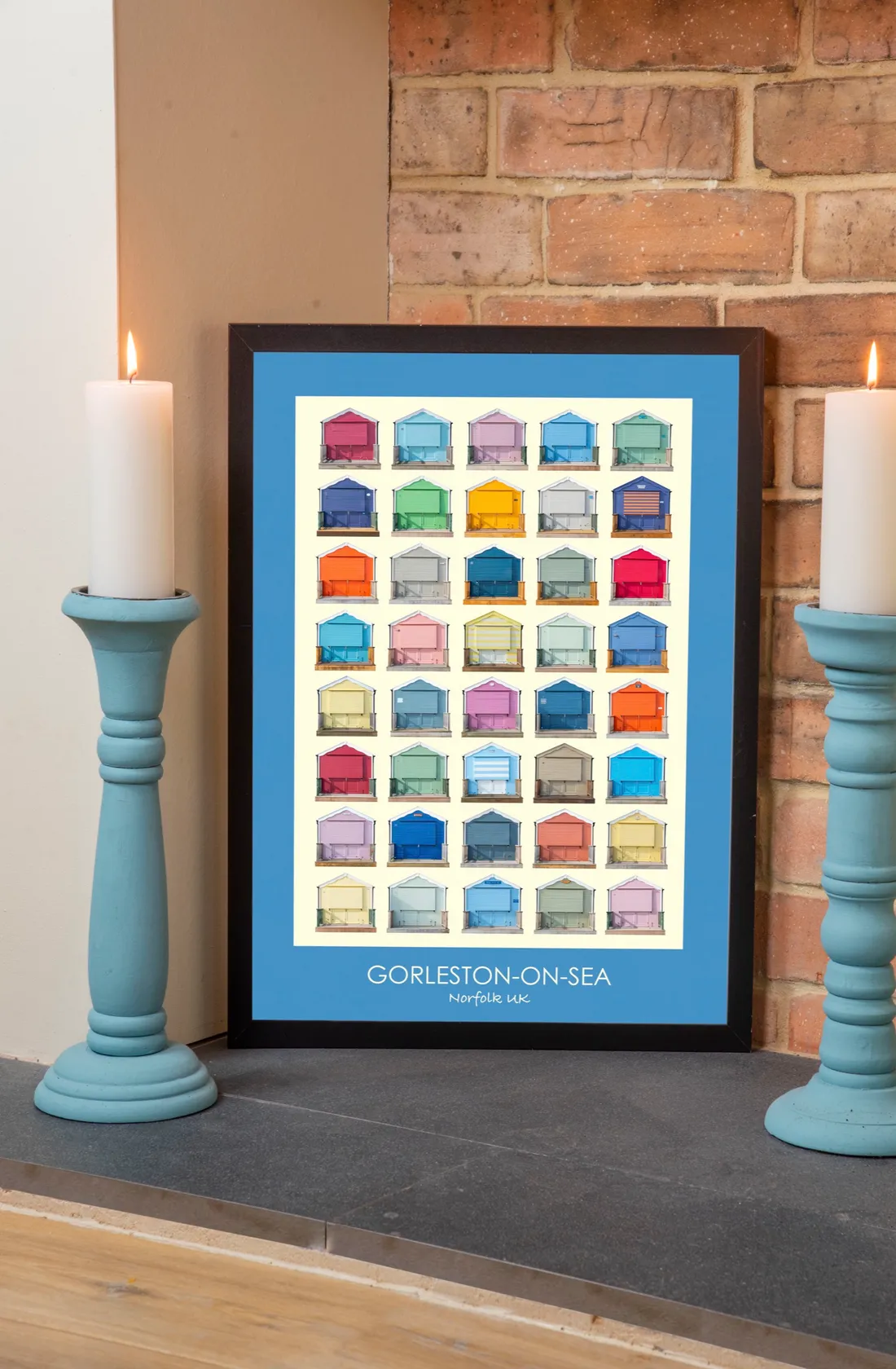 The Gorleston-on-Sea print with a True Blue border in a black frame, standing between two pale blue candlesticks with lit candles against a brick wall