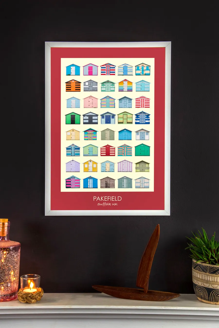 The Pakefield print with a spade red border in a white frame on a charcoal wall above a mantelpiece, with a candle, a wooden boat and a potted plant