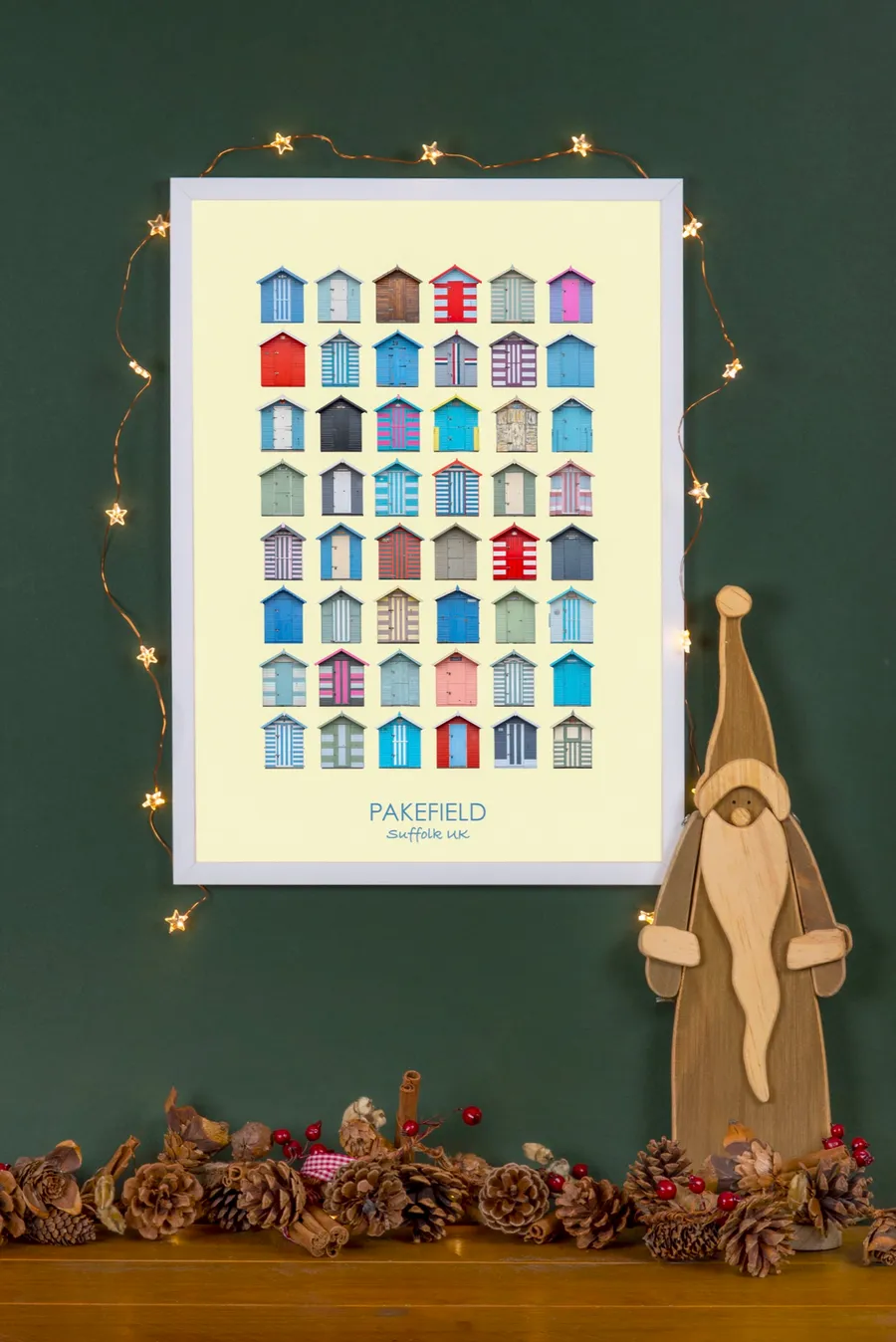 The Pakefield print in a white frame on a dark green wall, strung with fairy lights, beside a wooden Father Christmas figure and a garland of pine cones